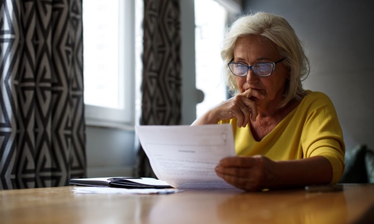 Egy szemüveges, idősebb nő egy iratot tanulmányoz egy szobában.