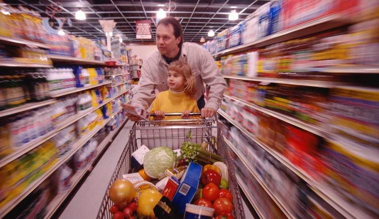 Egy apa és kislánya élelmiszerekkel telepakolt bevásárlókocsit tol a boltban.