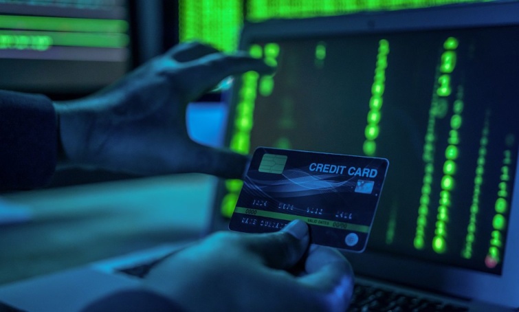 Egy laptop mögött sötétben ülő hacker egy bankkártyát tart a kezében.