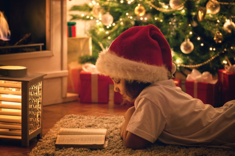 Egy mikulássapkás kislány a karácsonyfa alatt olvas.