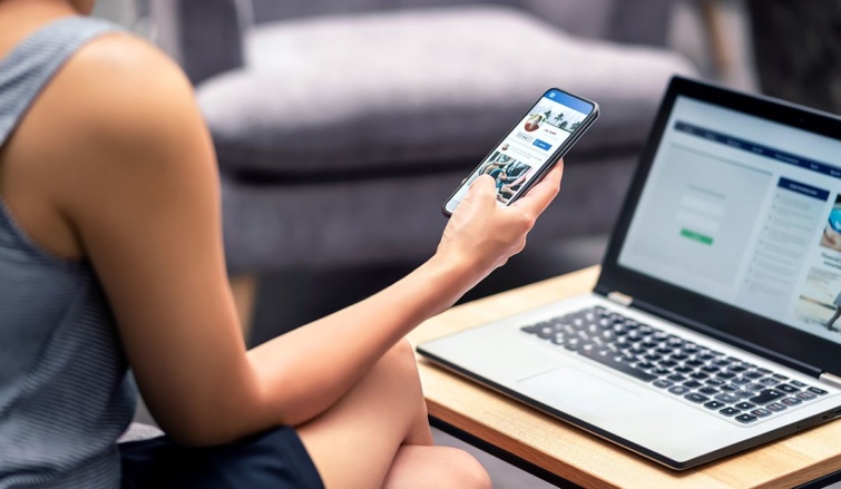 Egy nő egy mobiltelefont tart a kezében, amelynek kijelzőjén egy Facebook-profilt néz, a háttérben egy laptop.
