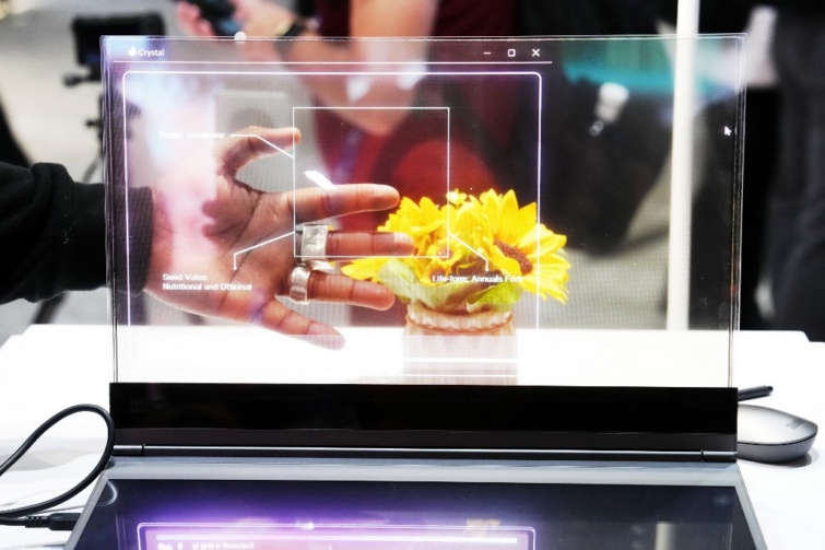 A Lenovo új, átlátszó kijelzővel ellátott laptopja látható a képen, fehér asztalon. A laptop előtt sárga színű cserepes virág.