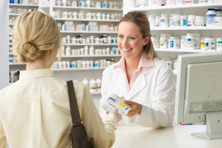 Gyógyszertári polc előtt mosolygós patikus hölgy nyújt át a vásárlónak egy gyógyszert.