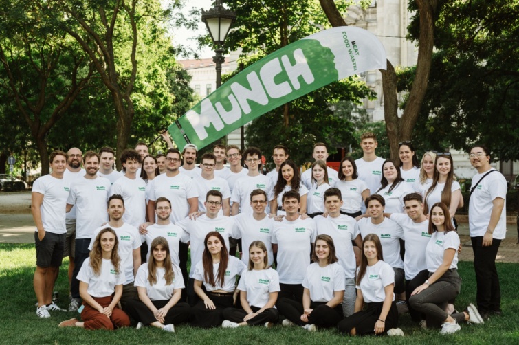 A Munch csapata egy zöld Munch feliratú molinó alatt, fák között, fehér polót visel mindenki