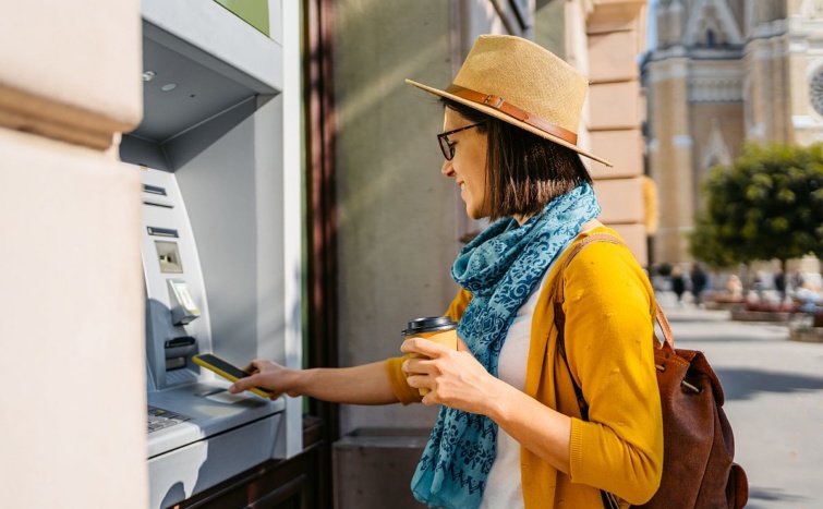 Egy fiatal nő a mobiltelefonját a bankautomatához érinti.