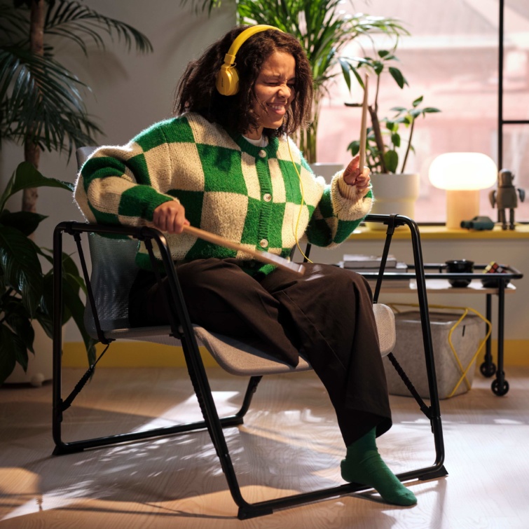 Mujer alegre con auriculares y una chaqueta verde y blanca sentada en una mecedora Ikea Hamer y jugando