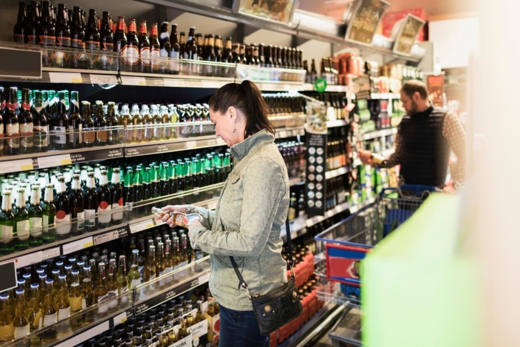 Egy boltban söröket nézeget egy barnahajú nő szürke kabátban