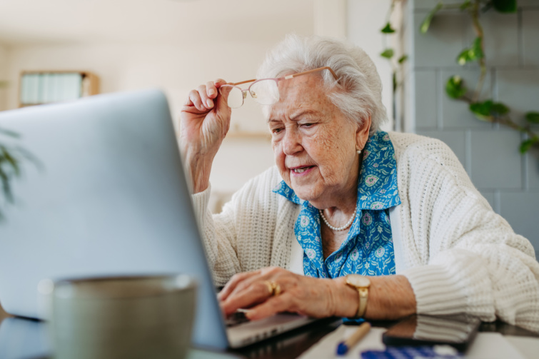 nyugdij Startlap Vásárlás Idős néni laptop előtt ül szemüveggel a kezében fehér pulóverben