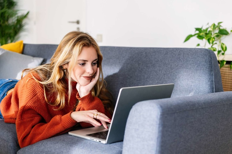 laptop Startlap Vásárlás Szőke nő narancssárga pulcsiban laptopozik a kanapén hasalva