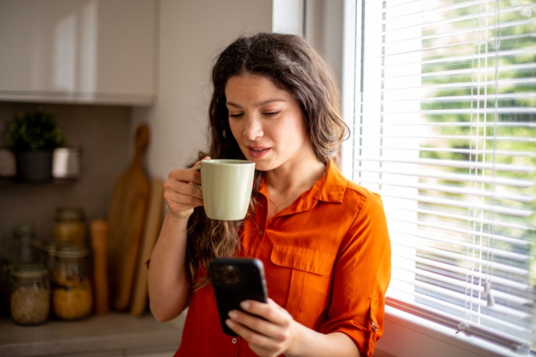 Fiatal felnőtt nő kávézik, miközben a telefonját nézegeti a konyhában.