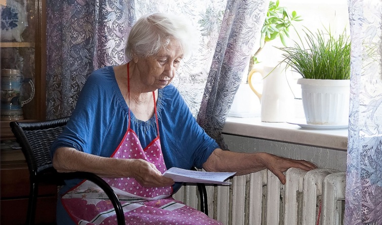 Egy idős nő egy iratot olvas a radiátor mellett.