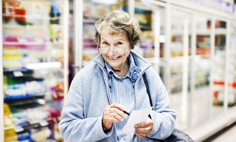 Senior woman in supermarket checks shopping list smiles delightedly Startlap Vásárlás Szemüveges idős nő tollal és bevásárlólistával a kezében a boltban belemosolyog a kamerába.