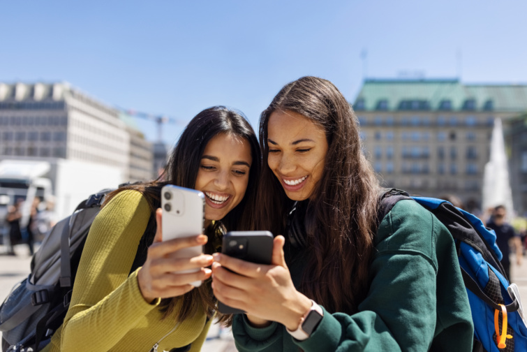 Két fiatal lány mosolyogva mutatja egymásnak a telefonját