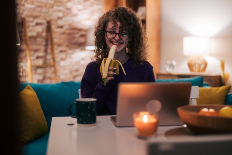 banan eves Startlap Vásárlás Fiatal lány banánt eszik egy laptop előtt ülve, körülötte gyertyák.