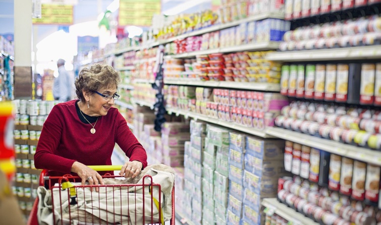 Idős, szemüveges nő bevásárlókocsit tol a boltban.