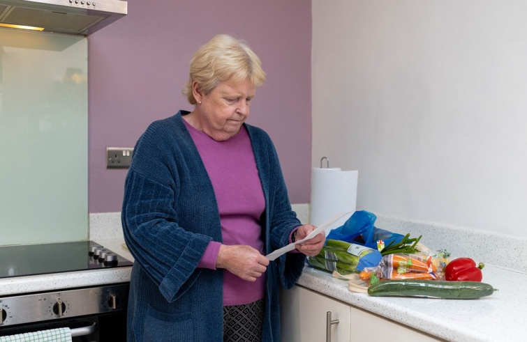 Egy idős nő egy nyugtát olvas, mellette zöldségáruk a konyhapulton.