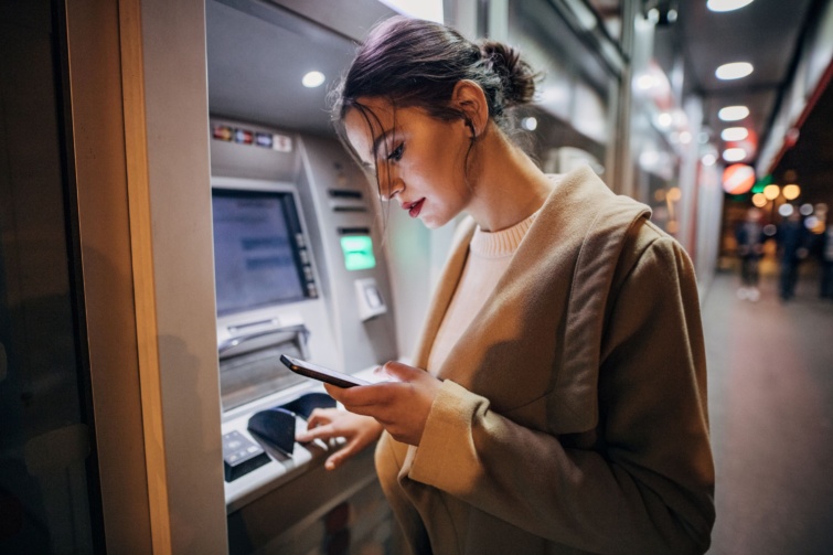 Fiatal lány a telefonját nyomkodja, miközben ATM-et használ.