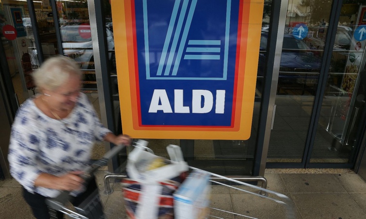 Egy elmosódott kontúrú vásárló egy bevásárlókocsival elghagyja az Aldi egyik boltját.