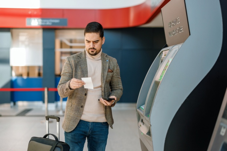 bankautomataferfiatm Startlap Vásárlás Üzletember készpénzt vesz fel egy ATM-ből a repülőtéren, miközben a telefonját nézi