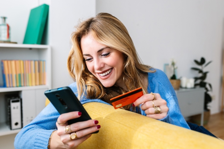 Boldog, fiatal szőke nő nyomkodja a telefonját kezében egy bankkártyával