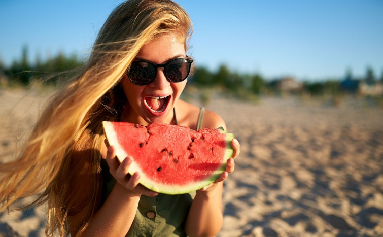 Happy young woman in glasses eating watermelon on the sandy beach on vacation Girl joyfully holding fresh watermelon skip in her hands and have fun with hair fluttering in the wind Youth lifestyle Startlap Vásárlás Fiatal, napszemüveges nő harapni készül egy görögdinnyeszeletből.