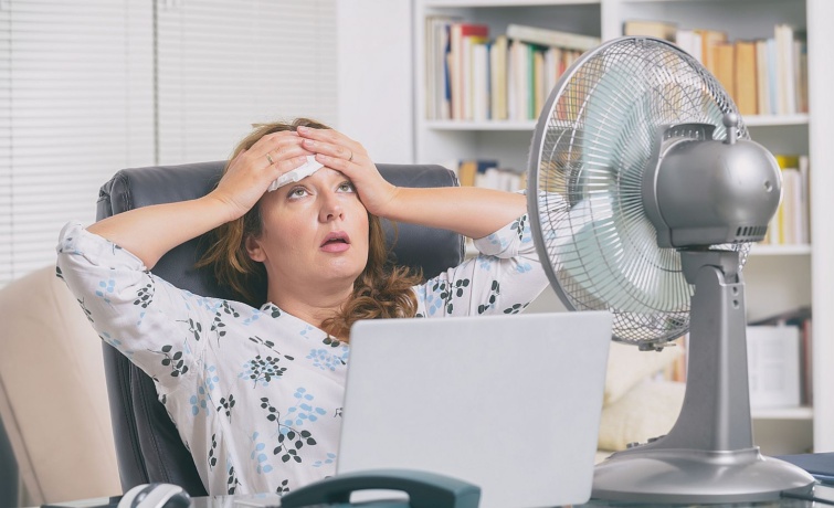 Fiatal nő szenved a hőségtől egy irodában, előtte laptop és ventilátor.