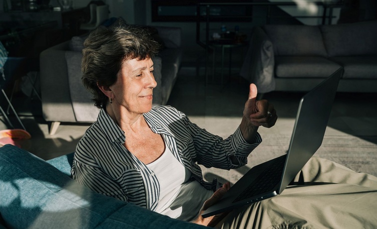 An older woman relaxes in a blue armchair with a laptop beside a sunlit window overlooking a private pool and tiled rooftops creating a peaceful remoteliving scene Startlap Vásárlás Egy idős nő laptoppal az ölében egy szobában a napfényben felmutatja a hüvelykujját