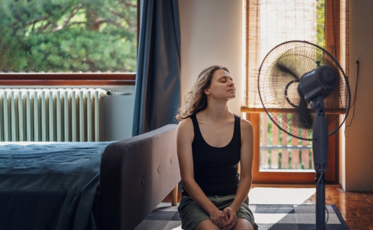 Csukott szemű hölgy hűti magát közvetlenül a ventilátor előtt ülve.