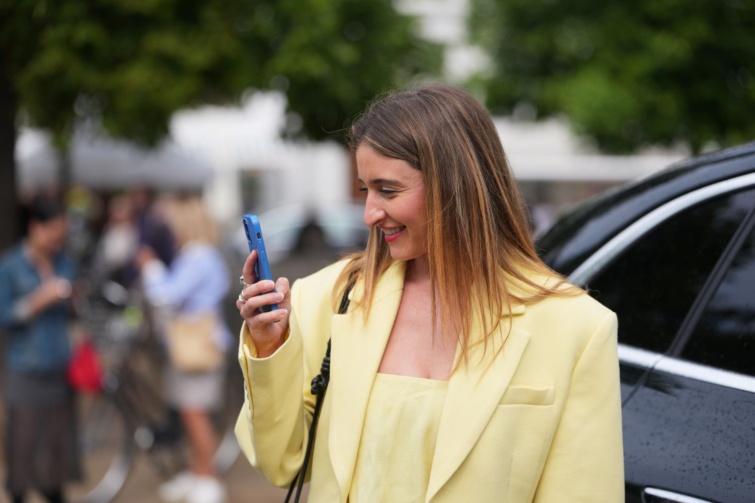 dáp Startlap Vásárlás Sárga zakót viselő hölgy mosolyogva vizsgálja a telefonját.