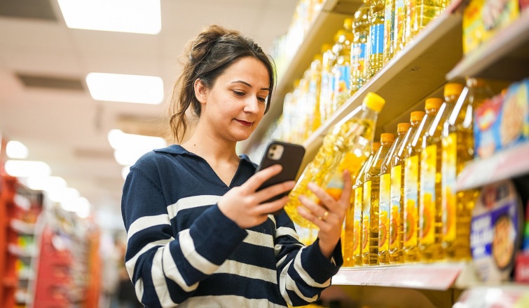 Young woman using smartphone to compare prices while shopping for cooking oil in supermarket Startlap Vásárlás Egy fiatal nő a mobiltelefonjával ellenőriz egy étolajterméket a boltban.