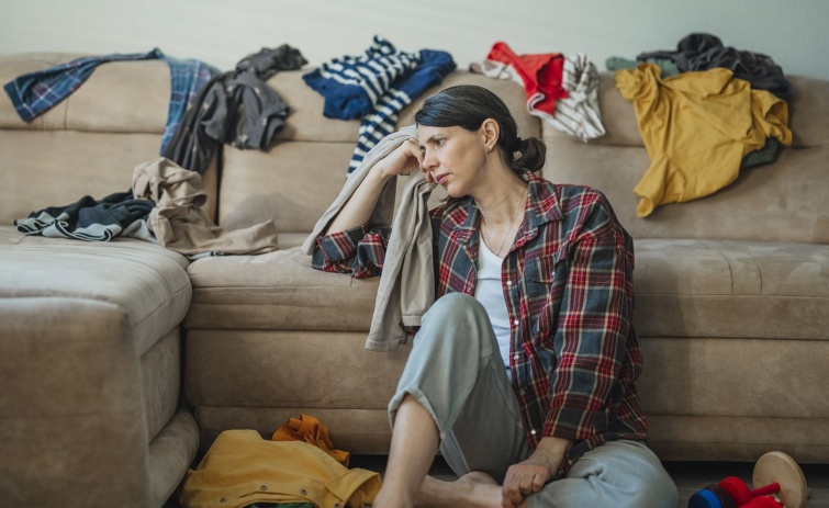 Tired woman sitting in living room with messy clothes on sofa Startlap Vásárlás Fáradt nő ül a kanapénak dőlve a földön szétszórt ruhák között.