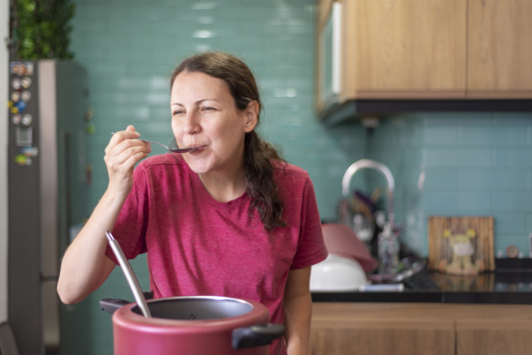 Fiatal nő mosolyogva ételt kostól egy edényből főzés közben