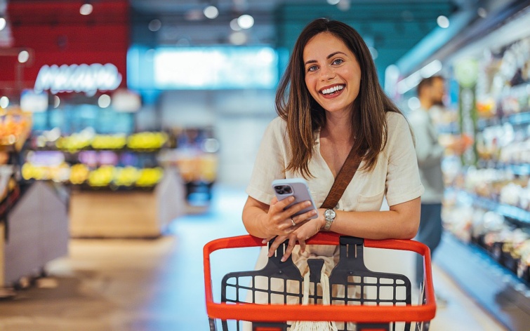 Portrait of happy woman buying groceries in supermarket and looking at camera Purchasing Goods with Smartphone at Grocery Store Female customer shopping with smartphone checklist Startlap Vásárlás Fiatal, mosolygó nő mobiltelefonnal a kezében bevásárlókocsira támaszkodik egy boltban.