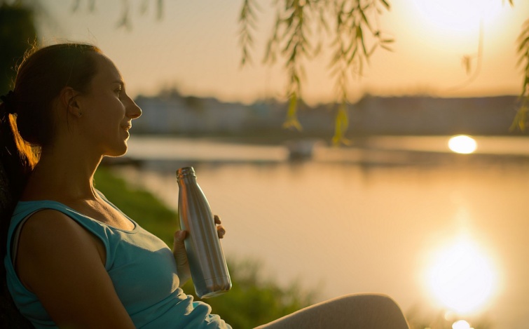 Woman with a reusable steel bottle of water in a park after running in sunset Startlap Vásárlás Fiatal nő egy tóparton, naplementében, kezében acélkulacs.