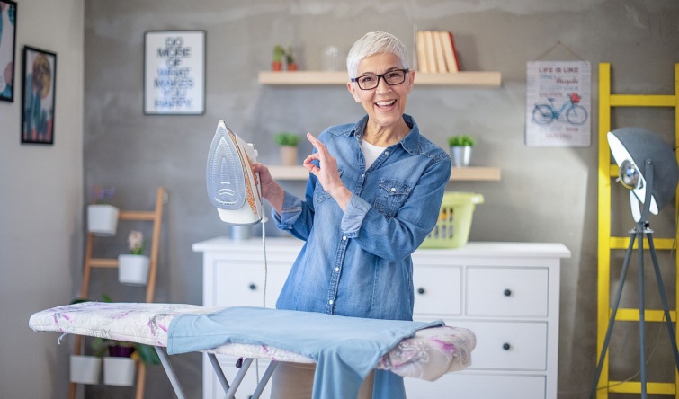 Beautiful mature senior woman with grey hair ironing her clothes during pandemic Covid19 Corona virus Startlap Vásárlás Idős, szemüveges nő az elégedettség jelét mutatja vasalás közben.