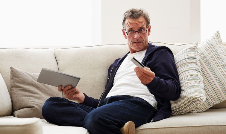 Frustrated senior man with digital tablet and credit card Startlap Vásárlás Egy szemüveges középkorú, tanácstalan férfi a kanapén ülve egy ik kezében tabletet, másikban bankkártyát tart.
