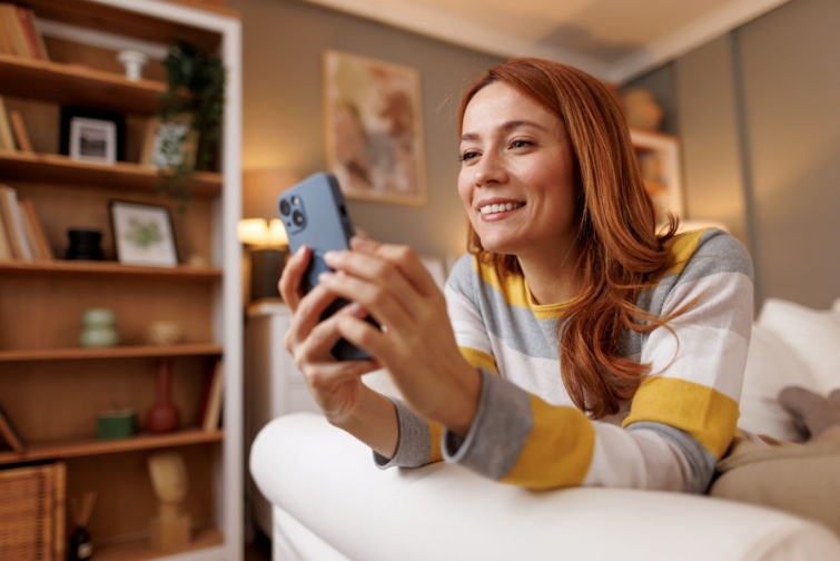 fiatalnotelefon Startlap Vásárlás Vidám, vörös hajú nő mobilozik, miközben egy kényelmes kanapén pihen a hangulatos nappaliban