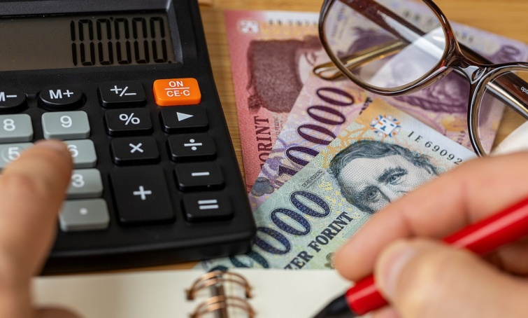 Forint bankjegyek és szemüveg az asztalon. Egy számológépen tartott ujj és egy tollat fogó kéz.