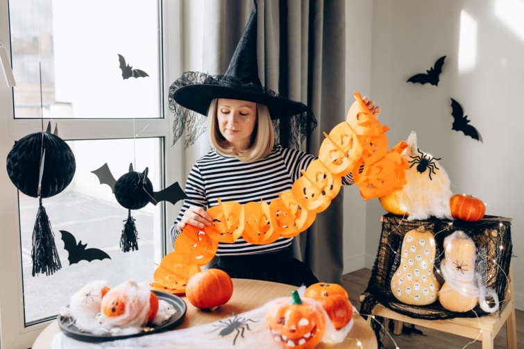 halloweendekoracio Startlap Vásárlás Boszorkányjelmezes szőke nő díszíti otthonát Halloweenre, papírfüzéreket akaszt fel egy otthonos lakásban